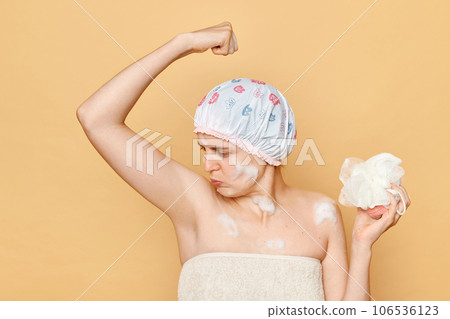 Young adult woman raised her arms smelling her armpit feeling unpleasant aromat frowning face being ready to take shower holding sponge wearing bath cap. 106536123