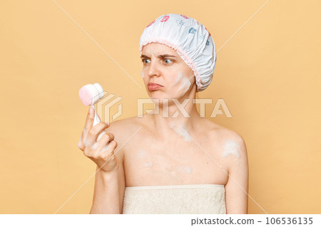 Confused puzzled woman wearing shower cap and wrapped in towel holding cleansing brush first time does't know how to use cosmetic tool standing isolated over beige background. Confused puzzled woman wearing shower cap and wrapped in towel holding cleansing brush first time does't know how to use cosmetic tool standing isolated over beige background. 106536135
