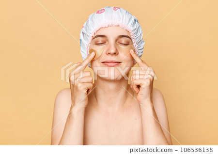 Perfect cosmetology treatment. Beauty patch application. Healthy skin care routine. Satisfied woman wearing shower cap with patches under closed eyes isolated over beige background Perfect cosmetology treatment. Beauty patch application. Healthy skin care routine. Satisfied woman wearing shower cap with patches under closed eyes isolated over beige background 106536138