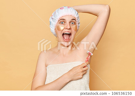 Excited female dressed wearing bath cap holding in hand razor shaver blade making depiliation armpit posing isolated on beige background. 106536150