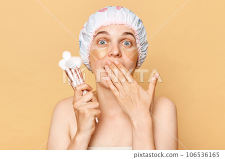 Astonished surprised woman doing makeup holding powder brushes posing with patches under eyes isolated over beige background covering mouth with palm. Astonished surprised woman doing makeup holding powder brushes posing with patches under eyes isolated over beige background covering mouth with palm. 106536165