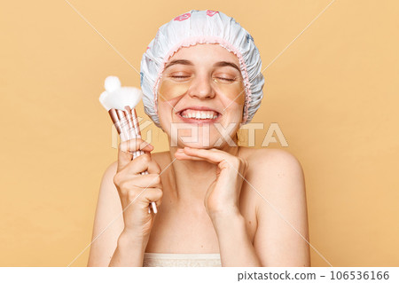 Smiling happy positive caucasian woman with shower cap applies hydrogel patches for skin treatment holds cosmetic brushes for applying makeup isolated over beige background. Smiling happy positive caucasian woman with shower cap applies hydrogel patches for skin treatment holds cosmetic brushes for applying makeup isolated over beige background. 106536166