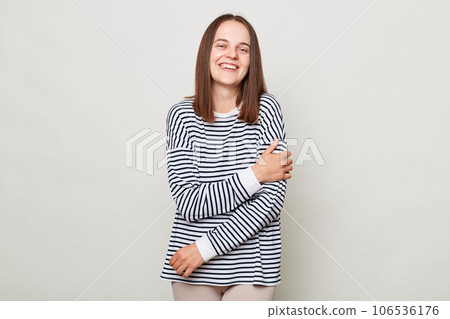 Pretty smiling happy woman with brown hair wearing striped shirt standing isolated over gray background looking at camera laughing expressing positive emotions. 106536176