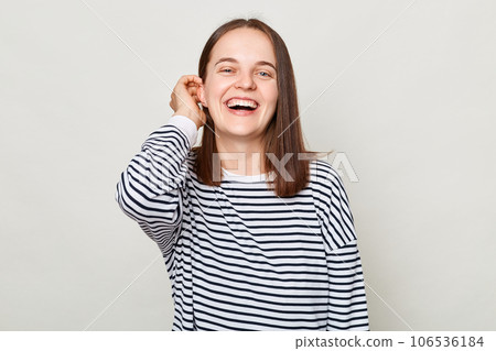 Beautiful optimistic woman with brown hair wearing striped shirt standing isolated over gray background being in good mood flirting keeps hand on hair. 106536184