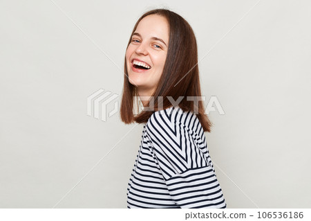Attractive positive joyful woman with brown hair wearing striped shirt standing isolated over gray background looking at camera and laughing. 106536186