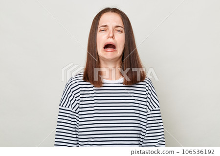 Unhappy sad brown haired woman wearing striped shirt posing isolated over gray background crying feeling despair hearing very bad news. Unhappy sad brown haired woman wearing striped shirt posing isolated over gray background crying feeling despair hearing very bad news. 106536192