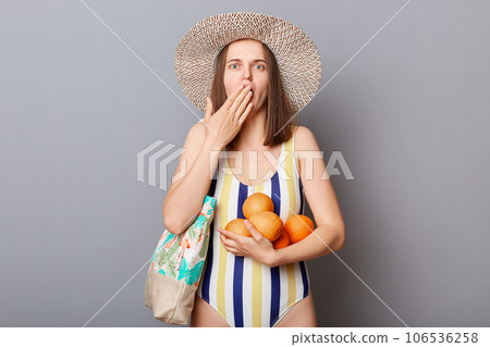 Shocked scared woman wearing striped swimsuit and straw hat looking at camera with big eyes covering mouth with hands isolated gray background, holding oranges. Shocked scared woman wearing striped swimsuit and straw hat looking at camera with big eyes covering mouth with hands isolated gray background, holding oranges. 106536258
