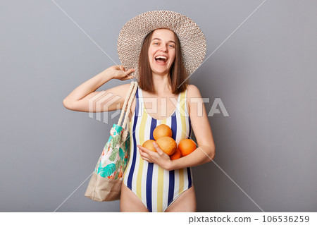Summer traveling. Smiling laughing young adult woman wearing striped swimsuit and straw hat holding fresh fruit enjoying resting having vacation isolated gray background Summer traveling. Smiling laughing young adult woman wearing striped swimsuit and straw hat holding fresh fruit enjoying resting having vacation isolated gray background 106536259