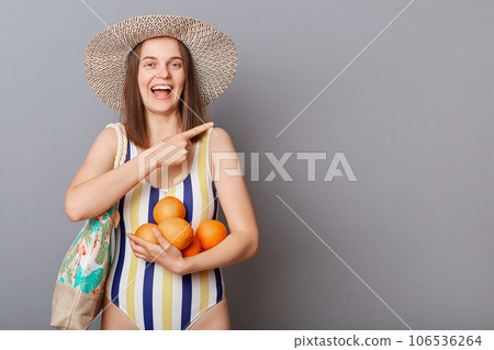 Smiling delighted woman wearing striped colorful swimwear standing with bag and oranges isolated over gray background pointing aside at copy space for promotional text. Smiling delighted woman wearing striped colorful swimwear standing with bag and oranges isolated over gray background pointing aside at copy space for promotional text. 106536264