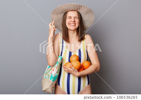 Hopeful smiling happy woman wearing striped colorful swimwear standing with bag and oranges isolated over gray background crossed fingers wishes to have good vacation. Hopeful smiling happy woman wearing striped colorful swimwear standing with bag and oranges isolated over gray background crossed fingers wishes to have good vacation. 106536265