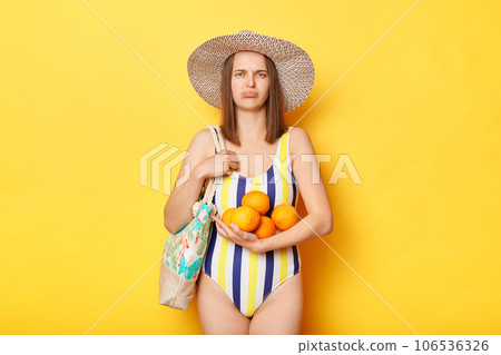 Disappointed woman wearing swimsuit isolated on yellow background holding fruit looking at camera with offended facial expression being sad. 106536326