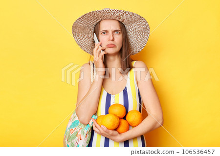 Sad upset woman on resort dressed in striped swimwear and straw hat posing isolated over yellow background holding oranges in hands and ta,king on mobile phone. Sad upset woman on resort dressed in striped swimwear and straw hat posing isolated over yellow background holding oranges in hands and ta,king on mobile phone. 106536457