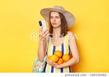 Sad upset displeased woman wearing striped swimming suit and hat isolated on yellow background holding fruit using mobile phone looking at smart phone screen with unhappy expression. 106536460