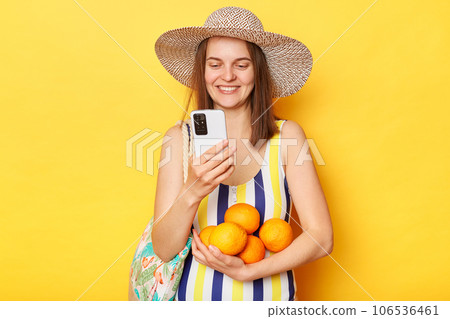 Joyful happy smiling woman wearing striped swimsuit and straw hat isolated yellow background holding mobile phone and fresh fruit checking social networks on her vacation. Joyful happy smiling woman wearing striped swimsuit and straw hat isolated yellow background holding mobile phone and fresh fruit checking social networks on her vacation. 106536461