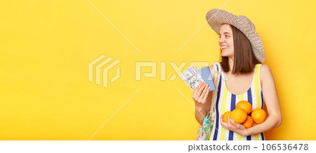 Satisfied delighted woman wearing striped swimsuit and straw hat isolated yellow background looking away at advertisement area holding documents and money. 106536478