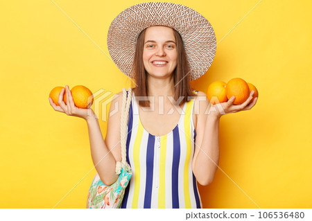 Smiling cheerful woman wearing striped one-piece swimsuit and straw hat posing isolated over yellow background standing with fresh oraganic fruit resting on vacation. 106536480