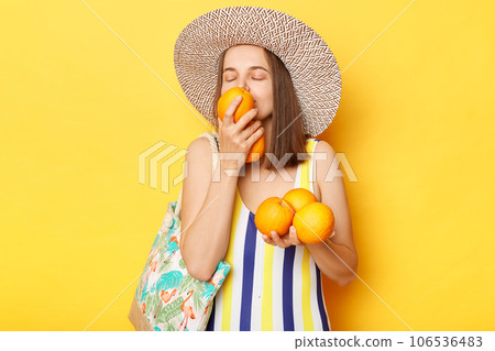 Satisfied delighted woman wearing striped one-piece swimsuit and straw hat posing isolated over yellow background standing smelling oraganic oranges resting on tropical resort. 106536483