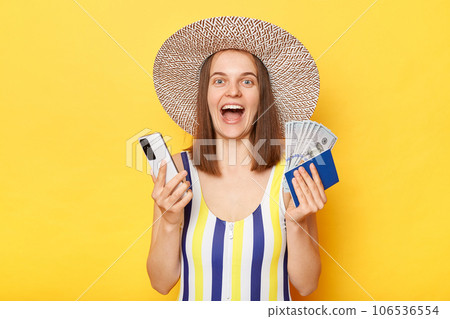 Overjoyed woman tourist holding cellphone tickets passport have excellent journey wearing striped swimming suit posing isolated over yellow background. 106536554