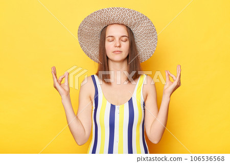 Calm woman on resort dressed in striped swimwear and straw hat posing isolated over yellow background practicing yoga meditating on resort keeps eyes closed. Calm woman on resort dressed in striped swimwear and straw hat posing isolated over yellow background practicing yoga meditating on resort keeps eyes closed. 106536568