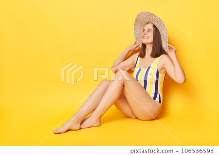 Enjoyable relaxed woman wearing one piece swimming suit posing isolated over yellow background sunbathing on beach expressing happiness enjoying summertime. 106536593