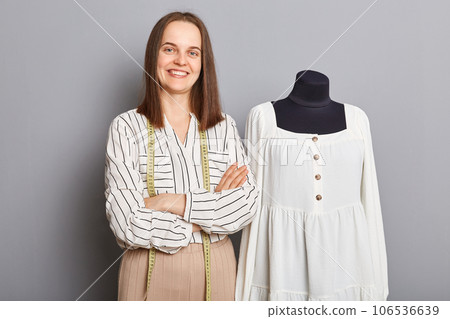 Tailor's creative work. Textile and fabric design. Clothing production process. Confident woman sewer standing with crossed arms near new dress on mannequin iisolated over gray background. Tailor's creative work. Textile and fabric design. Clothing production process. Confident woman sewer standing with crossed arms near new dress on mannequin iisolated over gray background. 106536639
