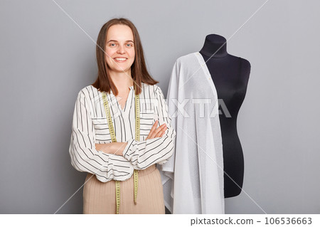 Attractive confident seamstress with measure tape on her neck standing with folded hands looking at camera creating new dress posing near menikin with white cloth isolated over gray background. 106536663