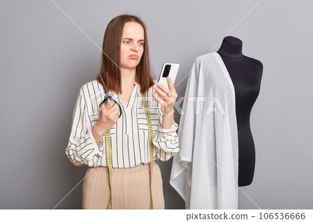 Sad upset professional female dressmaker expressing sorrow poising isolated over gray background using mobile phone checking orders in internet, has no work. 106536666