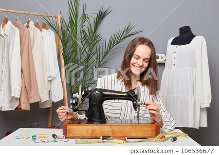 Caucasian young professional successful female designer at her workplace, dressmaker preparing sewing machine for sew clothes fabric in tailor workshop. Caucasian young professional successful female designer at her workplace, dressmaker preparing sewing machine for sew clothes fabric in tailor workshop. 106536677