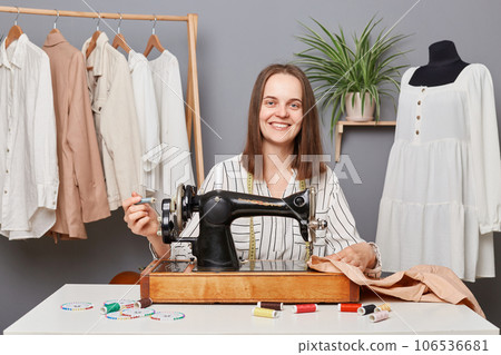 Successful caucasian female dressmaker businesswoman working on sewing machine in her own tailor workshop, looking at camera with happy facial expression. Successful caucasian female dressmaker businesswoman working on sewing machine in her own tailor workshop, looking at camera with happy facial expression. 106536681