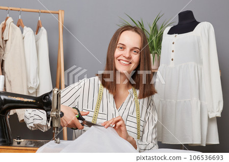 Pretty caucasian woman seamstress cutting textile for new dress in atelier, process of creating stylish clothing, female sewer working, looking at camera with happy expression. Pretty caucasian woman seamstress cutting textile for new dress in atelier, process of creating stylish clothing, female sewer working, looking at camera with happy expression. 106536693