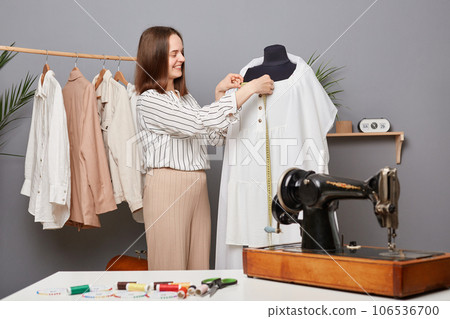 Satisfied professional seamstress measuring dress collar with tape during sewing process in atelier, enjoying working process, making handmade outfit in dressmaking workshop. Satisfied professional seamstress measuring dress collar with tape during sewing process in atelier, enjoying working process, making handmade outfit in dressmaking workshop. 106536700