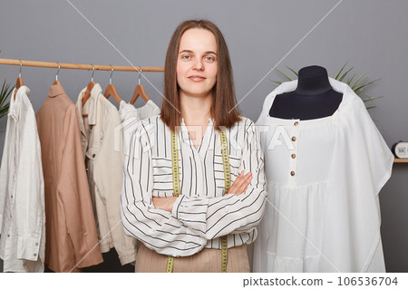 Confident successful dressmaker woman in atelier among fashion clothes collection, working in sewing workshop, dressmaking, posing with measure tape over neck, folded hands. 106536704