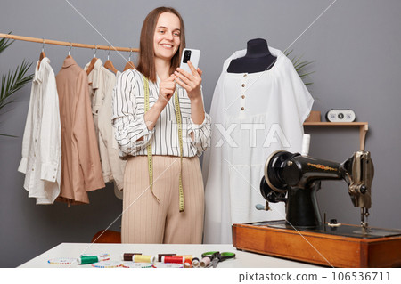 Portrait of beautiful woman seamstress holding phone in hand, tailor typing message on smartphone, searching patterns in internet, dressmaker working in workshop. Portrait of beautiful woman seamstress holding phone in hand, tailor typing message on smartphone, searching patterns in internet, dressmaker working in workshop. 106536711