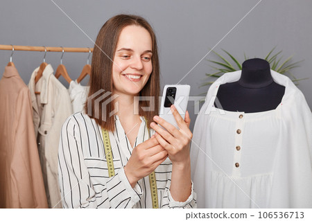 Smiling attractive young professional dressmaker using smartphone while working in atelier, searching new ideas in internet, browsing networks, fashion blog, sewing dresses for new collection. Smiling attractive young professional dressmaker using smartphone while working in atelier, searching new ideas in internet, browsing networks, fashion blog, sewing dresses for new collection. 106536713