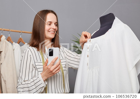 Delighted woman seamstress with phone in her hand working in atelier sewing white dress, smiling, enjoying her job in dressmaking workshop. 106536720