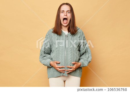 Shocked scared pregnant woman wearing knitted warm sweater standing isolated over beige background touching her belly screaming with fear feeling contractions. 106536768