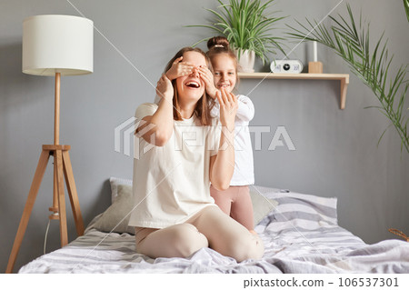 Happy funny woman and little daughter in bedroom mother and daughter sitting in bed having fun at home child covering mom's eyes playing hide and seek. 106537301