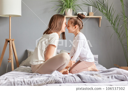 Side view portrait of brown haired woman and little girl, mother and daughter sitting in bed with closed eyes touching noses smiling playing together celebrating mother's day. 106537302