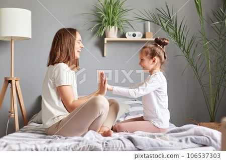 Side view portrait of mother and her daughter in bedroom at home, woman and little girl holding hands expressing happiness playing together with pleasure. 106537303