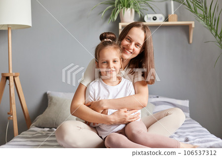 Home portrait of smiling woman and little female child mother and daughter sitting in bed hugging in bedroom looking at camera expressing happiness, mother's day congratulating. 106537312