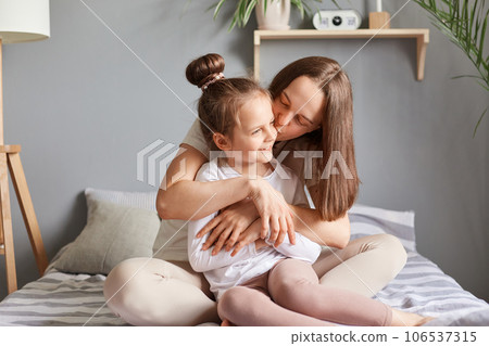 Charming little child girl congratulating mom with mother's day, daughter sitting in bed with mommy, gentle moment in morning, woman kissing her kid with love. 106537315