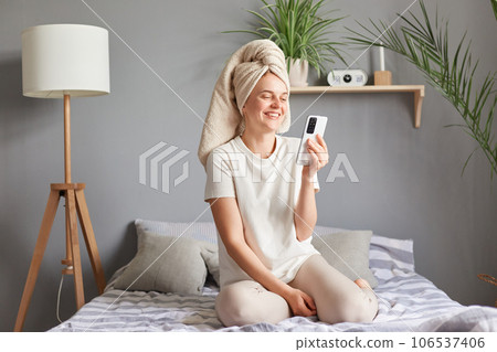 Morning beauty. Healthy relaxed woman sitting on bed using smartphone, having rest after bathroom, having video call laughing expressing positive emotions. Morning beauty. Healthy relaxed woman sitting on bed using smartphone, having rest after bathroom, having video call laughing expressing positive emotions. 106537406
