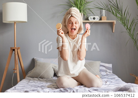 Extremely happy young Caucasian woman with towel on head doing skin care treatment on bed at home holding new sponges screaming with joyful expression. Extremely happy young Caucasian woman with towel on head doing skin care treatment on bed at home holding new sponges screaming with joyful expression. 106537412
