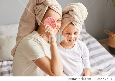 Happy child with mother sitting on bed in towels after morning shower having fun woman covering eye with cosmetic sponge family doing beauty procedures. 106537525