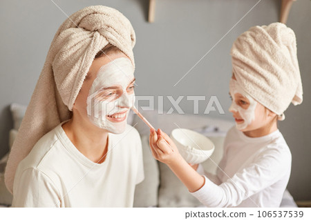 Indoor shot of smiling woman doing beauty procedures with her daughter, little kid applying mask to mommy face enjoying moment spending mother's day in spa. Indoor shot of smiling woman doing beauty procedures with her daughter, little kid applying mask to mommy face enjoying moment spending mother's day in spa. 106537539