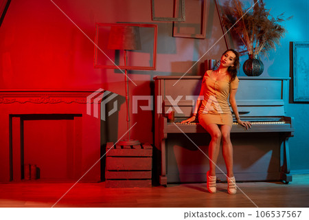 Young beautiful slim brunette girl with long curly hair in long beige dress with open shoulders stands near white grand piano in the room Young beautiful slim brunette girl with long curly hair in long beige dress with open shoulders stands near white grand piano in the room 106537567