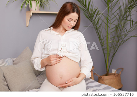Beautiful expectant mother sitting on bed, touching her bare tummy with affection, happily waiting for her baby's arrival, wearing white shirt looking at her belly. Beautiful expectant mother sitting on bed, touching her bare tummy with affection, happily waiting for her baby's arrival, wearing white shirt looking at her belly. 106537716