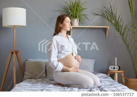 Beautiful gentle brown haired pregnant woman with bare belly wearing white clothing sitting in bed in light bedroom, touching her tummy, looking away, thinking about expecting baby. Beautiful gentle brown haired pregnant woman with bare belly wearing white clothing sitting in bed in light bedroom, touching her tummy, looking away, thinking about expecting baby. 106537717