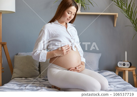 Beautiful pregnant woman with bare belly wearing white clothing touching and looking at her tummy and caring about her health sitting in bed in light bedroom. 106537719