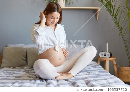 Winsome woman wearing white clothing sitting in bed in light bedroom, expectant lady touching belly and smiling, pregnant girl feeling baby kicks, enjoying last months pregnancy. 106537720